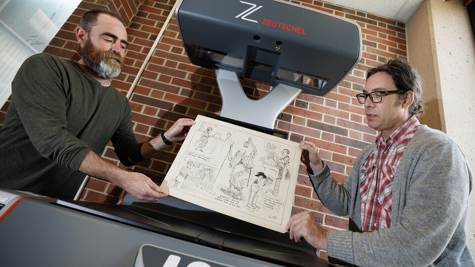 Richard Graham, Associate Professor of University Libraries, right, and John Wiese place an Oz Black cartoon onto the scanner bed to digitize the drawing. Graham and Wiese are building an online archive of editorial cartoons from Nebraska alumnus Oz Black. These cartoons are mostly from the 1920s and 1930s, and ran in Nebraska newspapers headquartered in Lincoln.