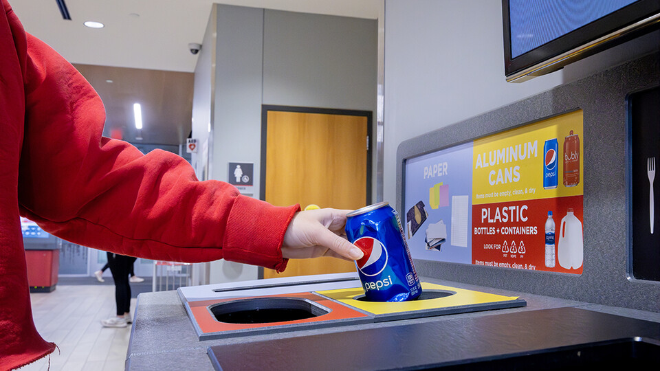 A hand places an aluminum can into an All in the Hall recycling station.