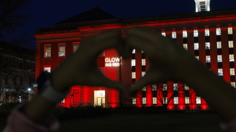 A student uses her hands to form a heart around the "Glow Big Red" words projected onto the south facade of Love Library.