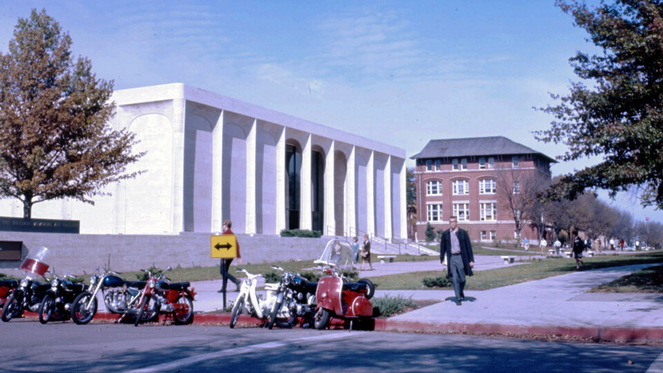 Students stroll under a blue sky along the sidewalk on the north side of 12th and R streets with Sheldon Museum of Art and the Geography Building in the background in this image from November 1965.