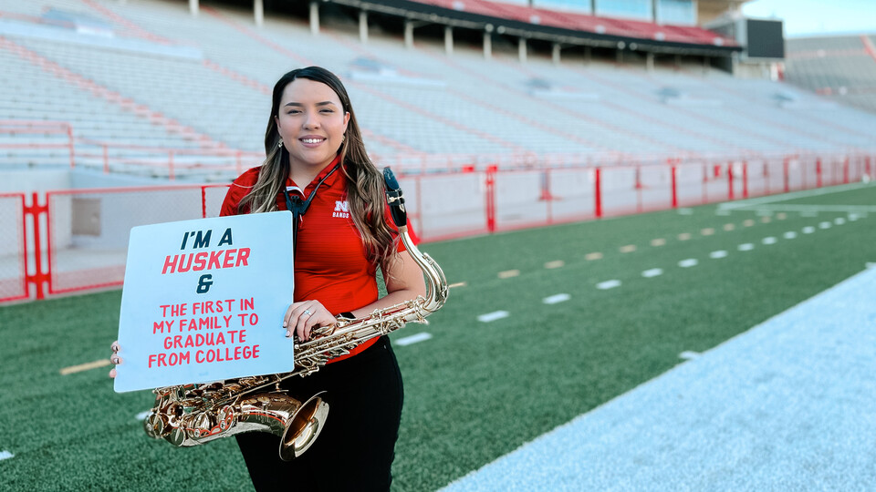 Aracely Acosta, a summer 2022 graduate, poses with her saxophone and an "I'm a Husker and..." sign while standing on the turf in Memorial Stadium.
