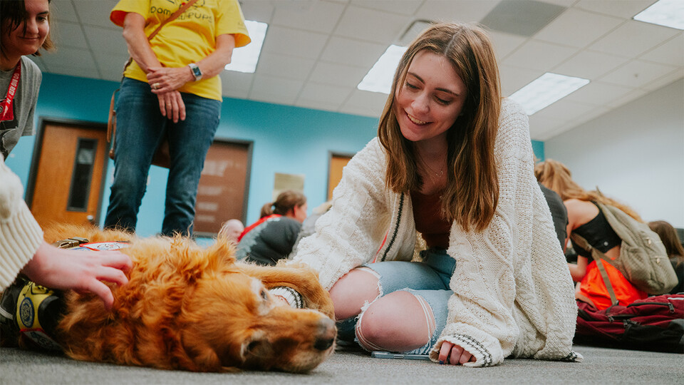 A Husker smiles while petting a dog that's lying on the floor