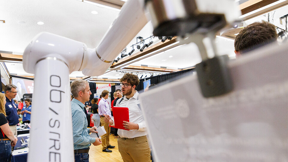 Framed by a robotic arm in the foreground, Leandro Castellanos Izaguirre chats with a potential employer at a career fair