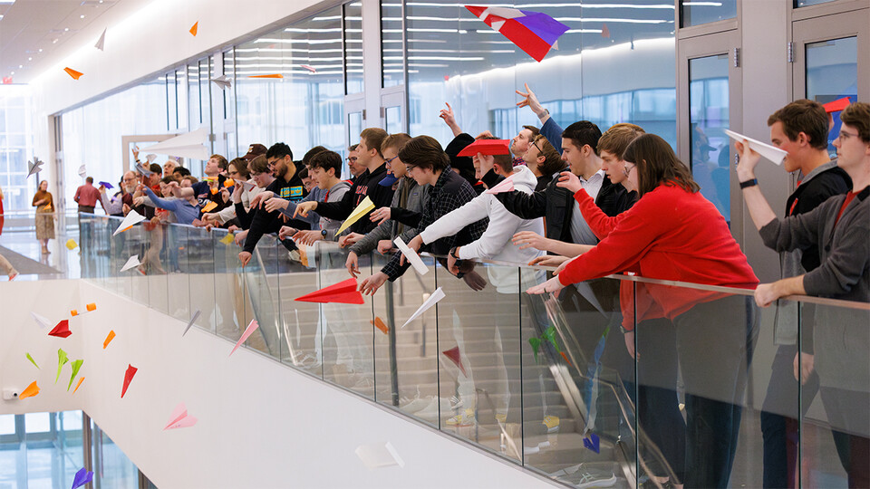 Standing in the open corridor of an upper level, Nebraska Engineering students, staff and faculty toss paper airplanes across the atrium of Kiewit Hall 
