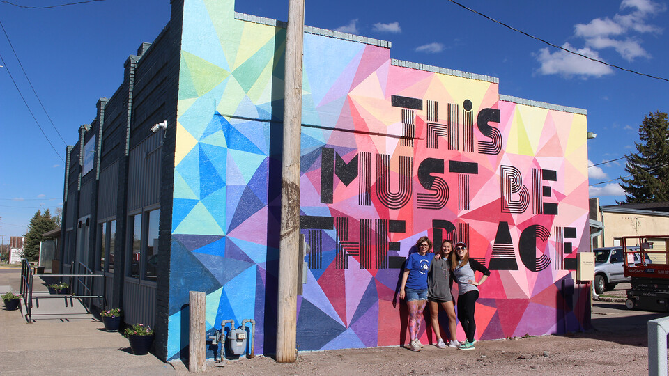 Three individuals standing in front of a colorful "This must be the place" mural in Alliance, Nebraska. Sandra Williams' outreach helped create the mural.