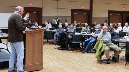 A man at a lectern speaks to about 20 people seated around tables.