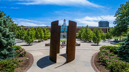 Three male students are framed by the "Greenpoint" sculpture as they walk west, with Love Library in the background.