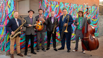 Six men with jazz instruments stand in front of a colorful mural on a building.