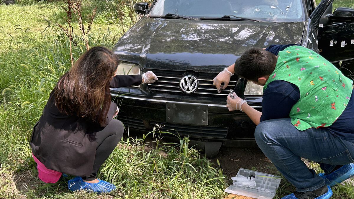 A man and woman examine insects on a car grille at an outdoor crime scene.