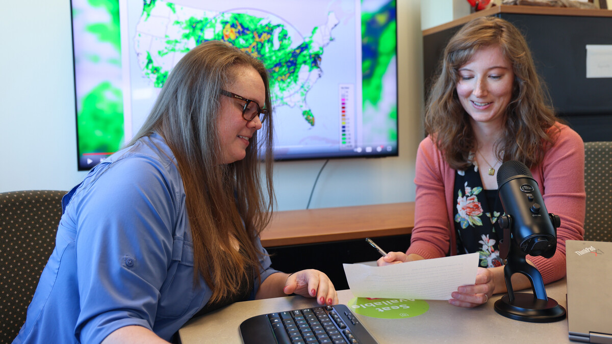 Lindsay Johnson (left) and Emily Case-Buskirk look at a piece of paper at a table with a microphone on it. In the background is a computer monitor with a map of the United States showing "NOAA Gridded 7-Day Precipitation."