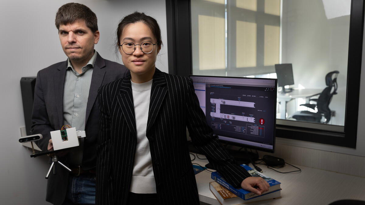 Iason Konstantzos (left) and Xiaoqi (Clare) Liu pose next to a computer and window. Through the window, a desk with a computer on it and an office chair are visible. Konstantzos holds a camera and sensor on a small tripod, and Liu rests her left arm on two textbooks.
