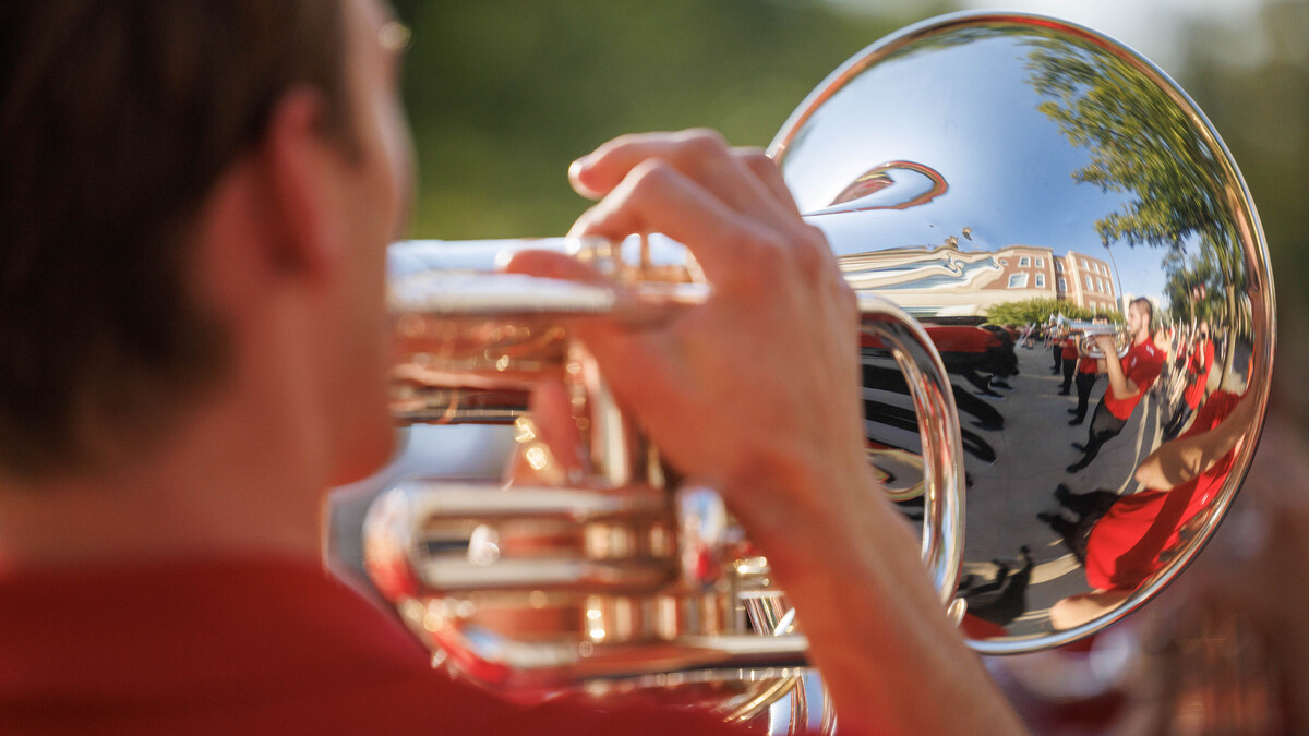 A male horn player plays, with other members of the Cornhusker Marching Band reflected in the bell of his instrument.
