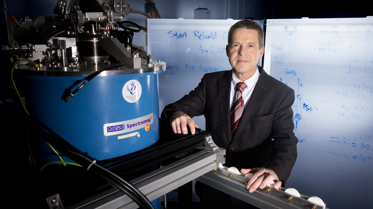 Mathias Schubert, J.A. Woollam Distinguished Professor of Engineering at the University of Nebraska–Lincoln, stands next to an Oxford Instruments Spectromag. He is wearing a dark suit, and behind him are two markerboards with writing on them.