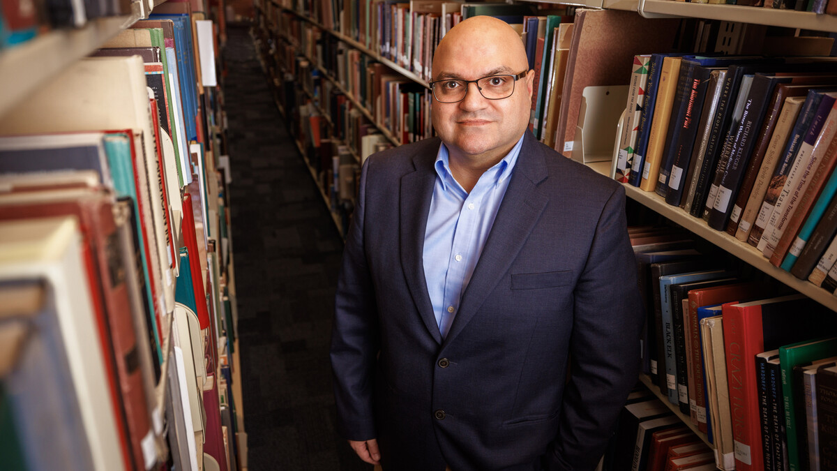 Bedross Der Matossian, Hymen Rosen Professor in Judaic Studies at the University of Nebraska–Lincoln, stands between rows of bookshelves in Love Library. He is wearing a dark blue sport coat over a light blue dress shirt and glasses.