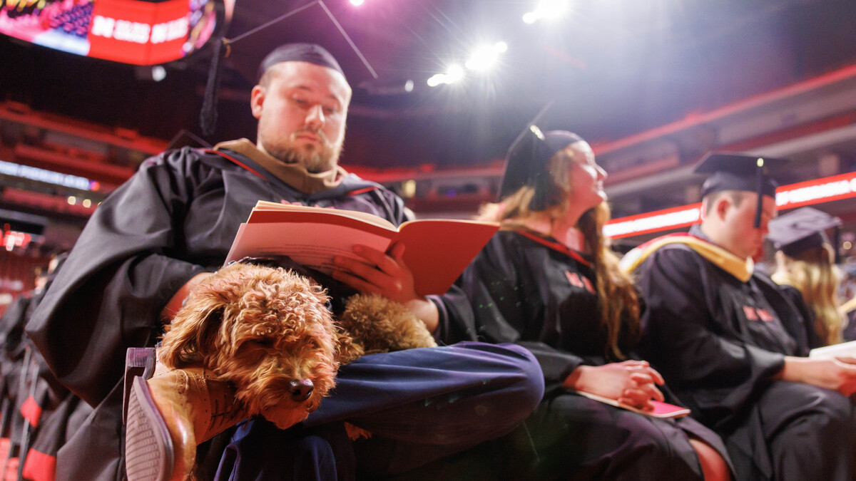 A shaggy brown dog rests on a graduate's lap as the graduation reads the program.