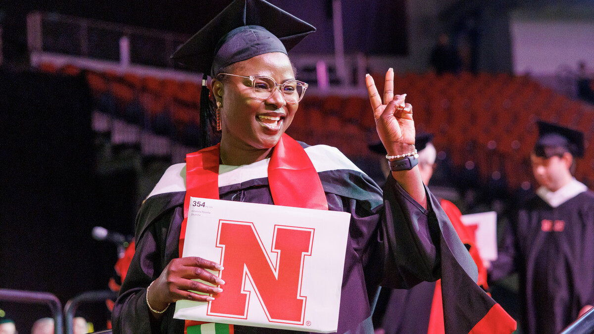 A female graduate smiles and displays a peace sign as she holds her newly earned diploma.