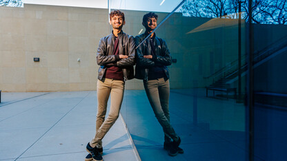 Aaryan Naik leans against Hawks Hall for a photo with his reflection in the glass of the building