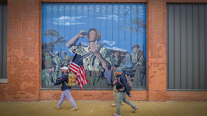 Nebraska alumni and veteran Pete Lass carries the American flag as he and Atlantic-area veterans Trevor Gipple (carrying game ball) and Ryan Graham walk past a mural on the side of the Coca-Cola Bottling Company in downtown Atlantic, Iowa, on the second day of the march.