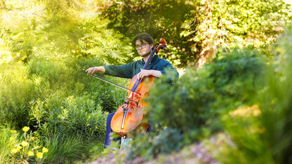 A student plays a cello outside in a greenspace on campus.