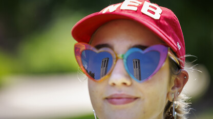 Student wearing a red "NEB" hat and wearing sunglasses with Mueller Tower reflected in heart-shaped lenses.
