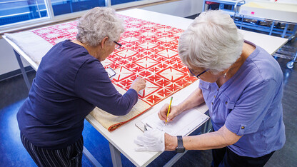 Two women lean over a red and white quilt spread over a table.