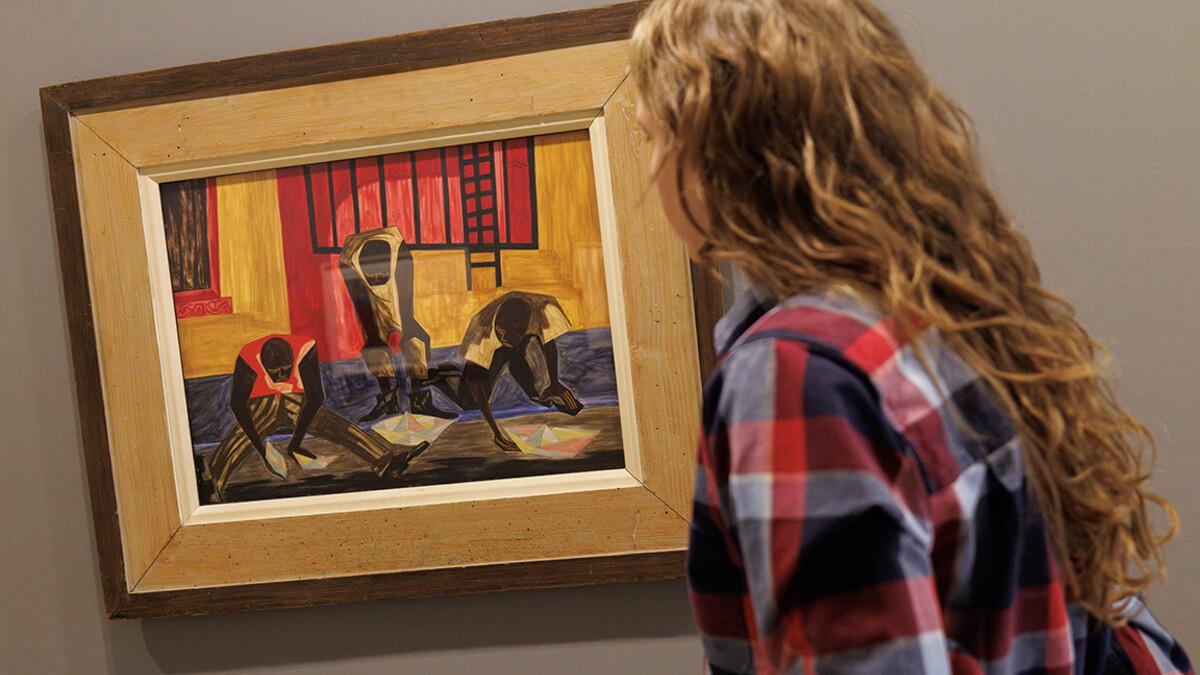 A person stands next to the framed painting "Paper Boats" by Jacob Lawrence