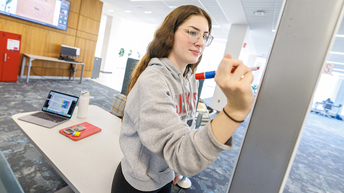 A white, brunette student with glasses, in a grey Nebraska sweatshirt, writes on a whiteboard in the library.