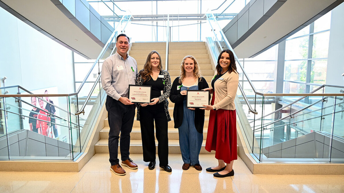 Four people holding two certificates smile for a photo in front of a white staircase. Right to left: a white man in black slacks and a. blue shirt; A white woman with long brown hair in a black shirt, slacks, and a grey cardigan; a white blonde woman in a blue jumpsuit and black cardigan; a white woman with long dark brown hair in a red skirt and cream cardigan.