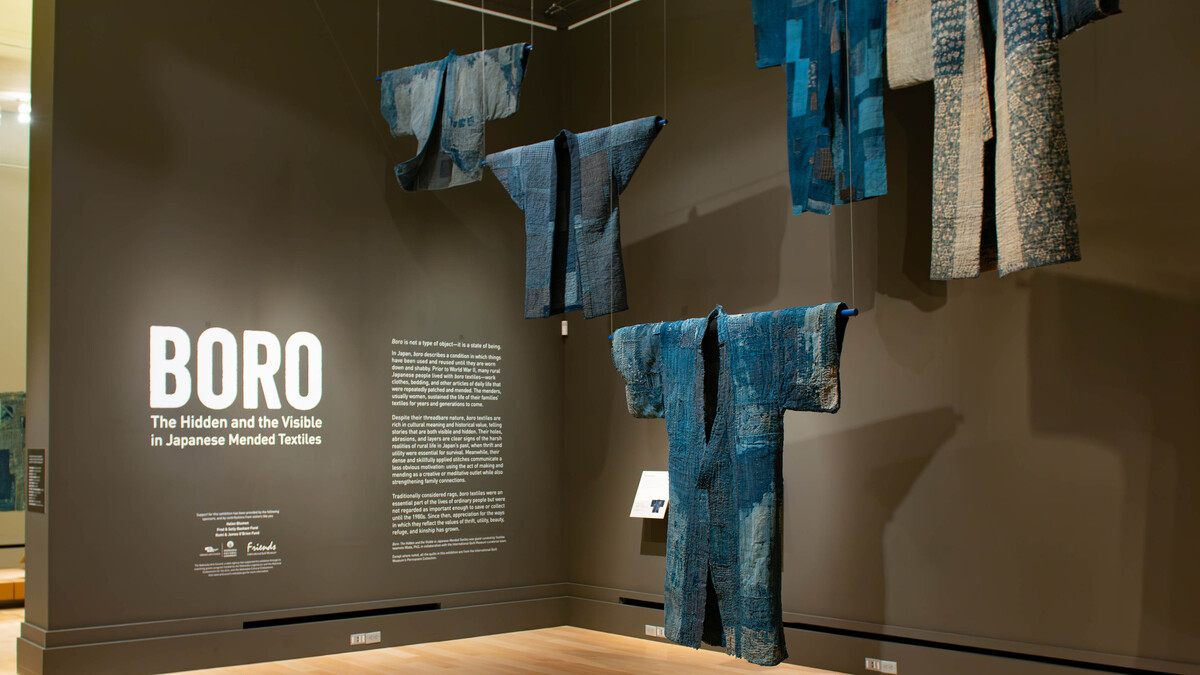 An art exhibition where dark blue or indigo quilts have been hunt up to resemble a sort of upside-down triangle, with text on the wall to its right, naming the exhibit BORO.