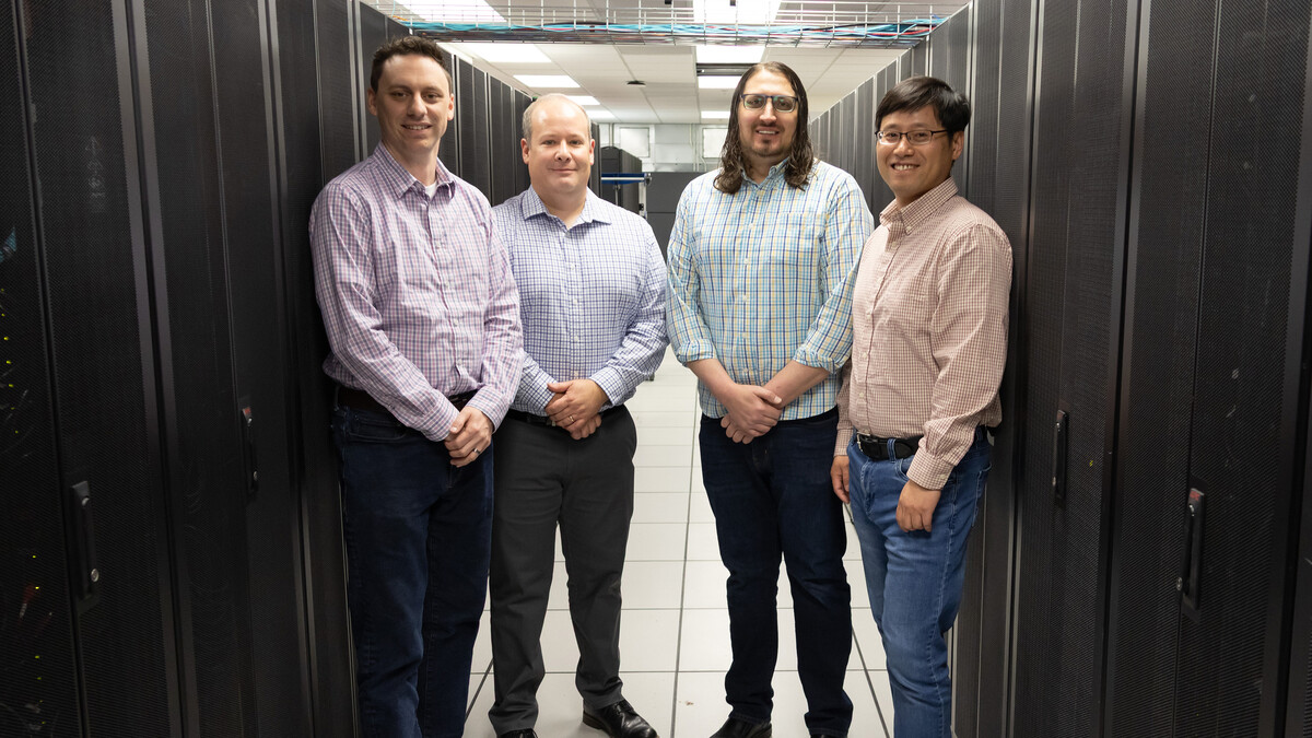 A white man with short brown hair and a purple button-up, white bald man in a blue button-up, white man with long dark hair, beard, and glasses in a turquoise button-up, and asian man with short brown hair in an orange button-up all stand together.