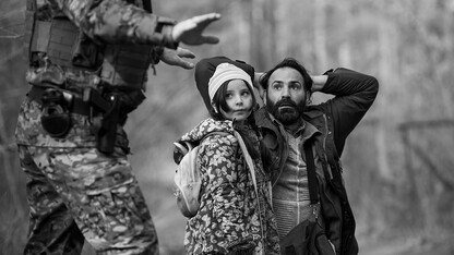 Black and white image of a young daughter standing next to her father, who is kneeling with hands behind his head after being confronted by a border guard.