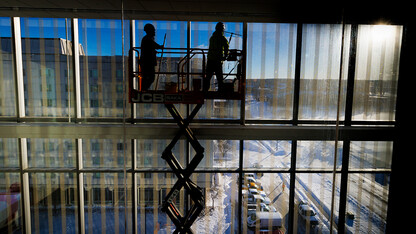 Two hard hat-clad workers atop a scissor lift clean a wall of windows in Kiewit Hall while silhouetted against a view of City Campus