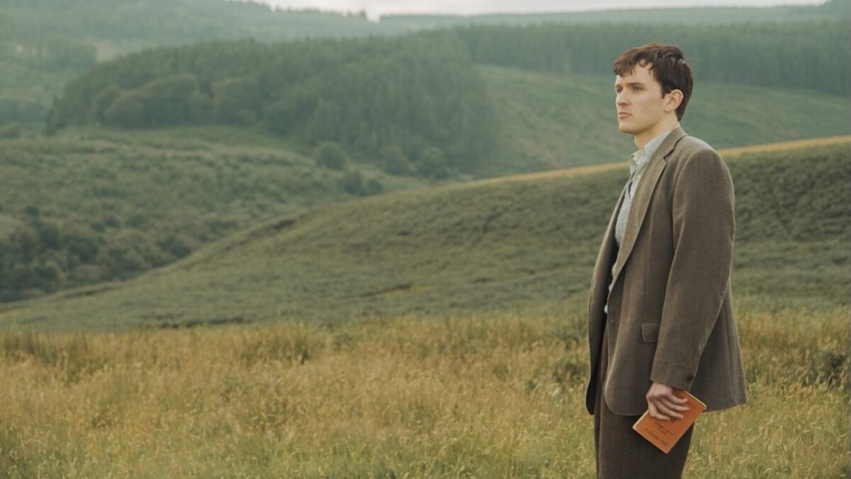 Harry Lawtey plays Richard Burton, a white man with dark short hair, standing in a green field in a brown suit.
