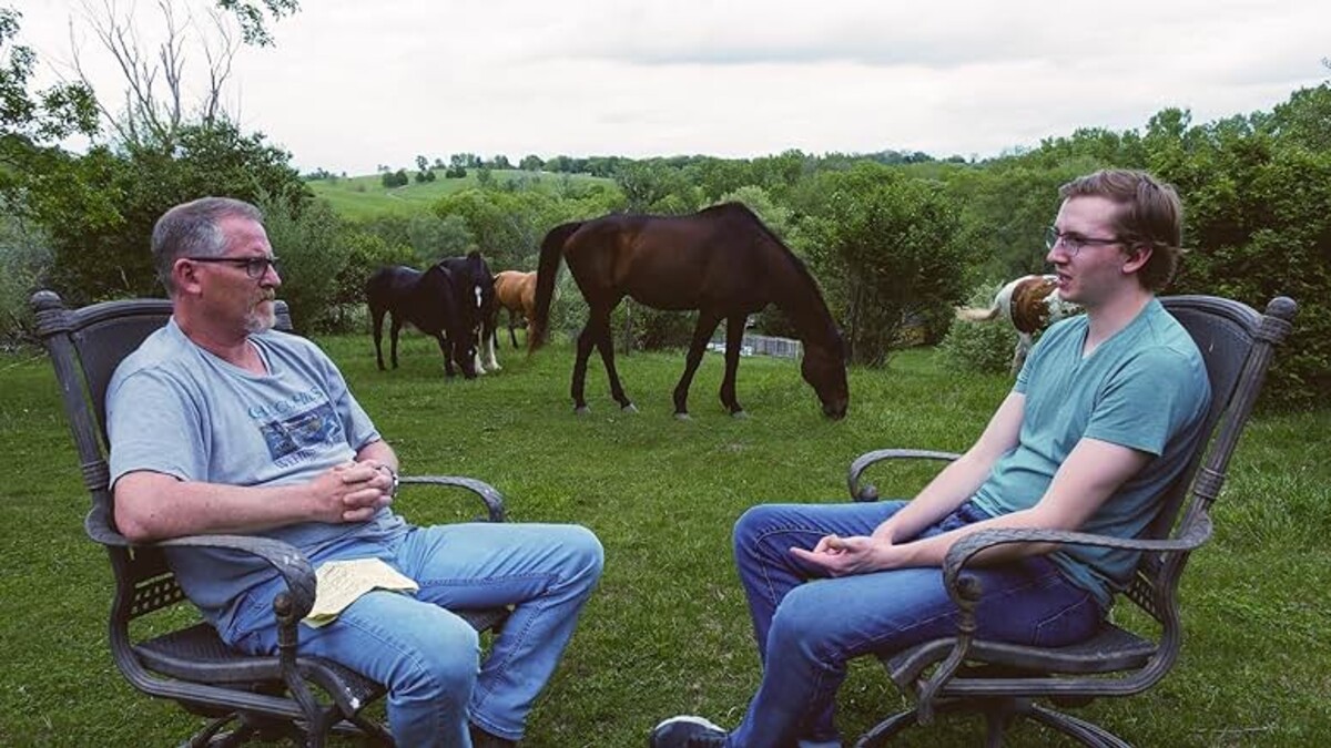 An older white man and younger white man, both in a t-shirt, jeans and glasses, sit across from each other in a green field outside, two horses grazing behind them.