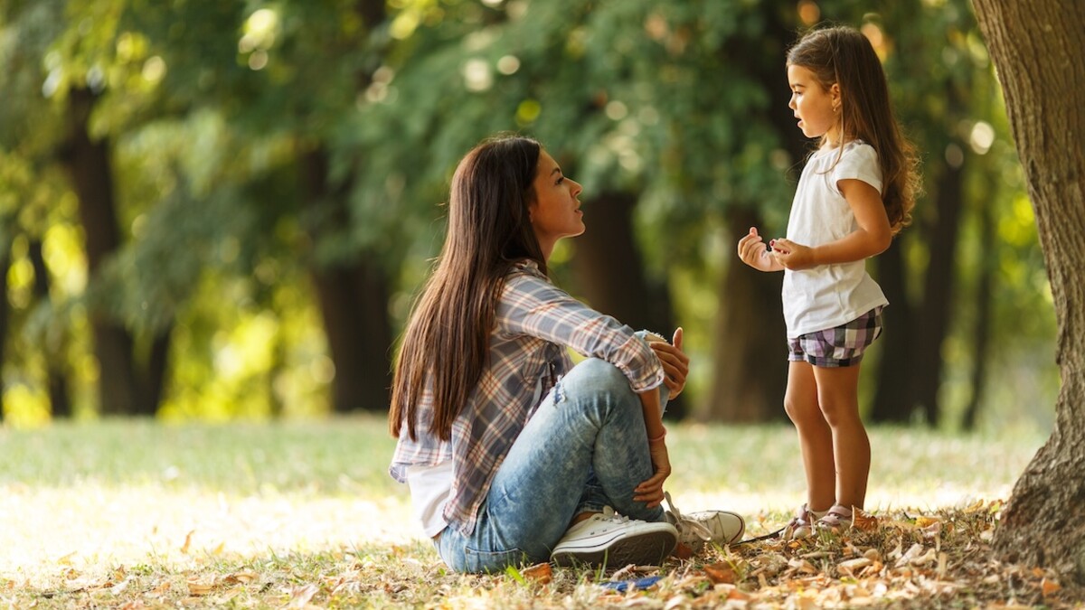 A mother sits next to a standing child under the canopy of a large tree.