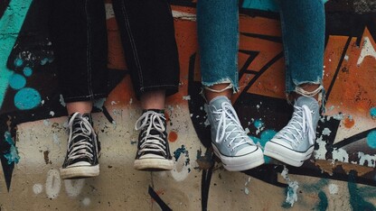 Photo by Aedrian Salazar on Unsplash - image shows the legs of two young people wearing blue jeans and sneakers, sitting on the edge of a painted skatepark wall.