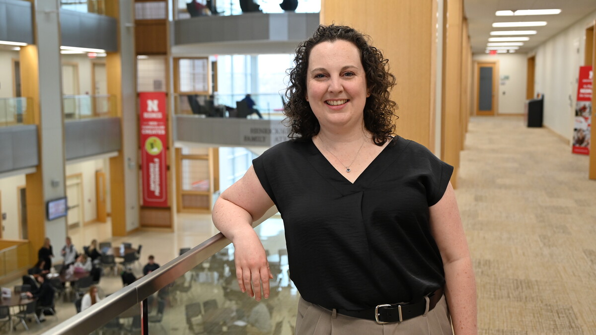 Rebecca Jack, a white woman with long, curly dark hair stands in a black blouse and tan trousers in the College of Business.