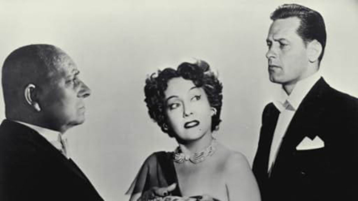 A woman with short, dark hair stands between two men, one short and bald on the left, one tall with cleanly done hair on the right in a black and white photo.