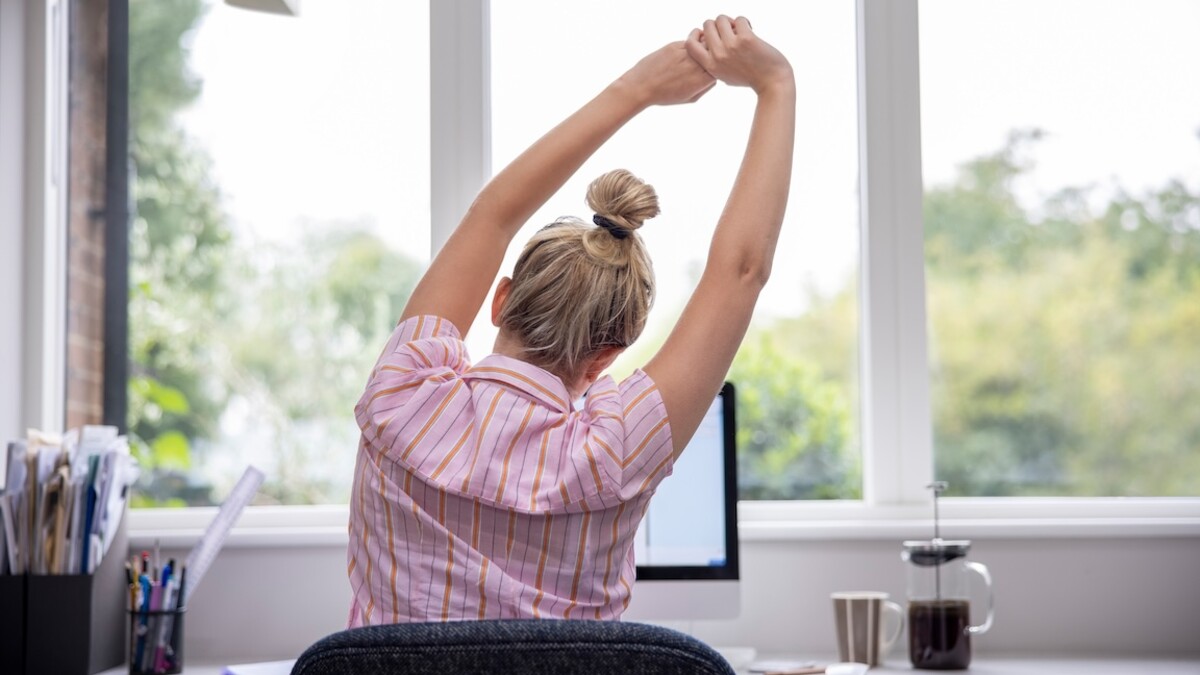 A woman stretches her arms into the air while seated at a desk.
