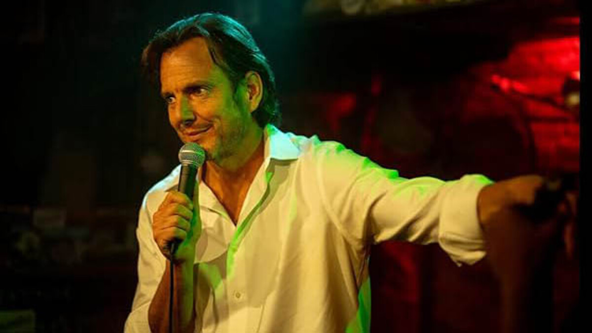 Actor Will Arnett, a white man with shoulder-length brown hair, stands holding a microphone in a white button-up shirt on stage.