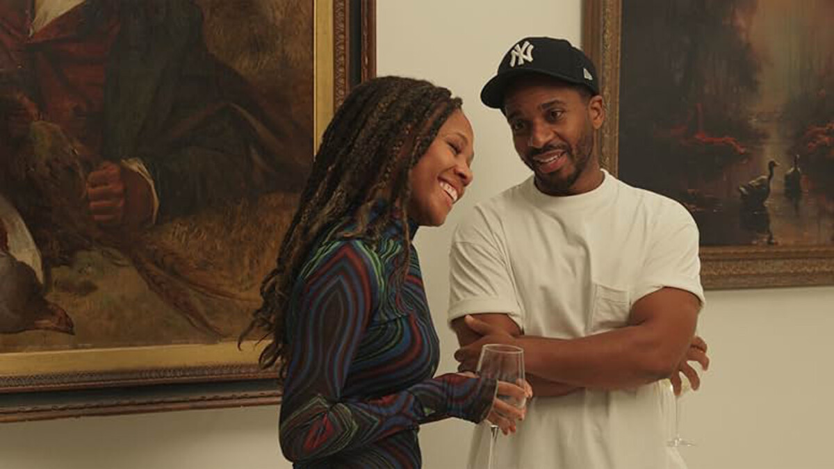 A black man and woman stand together by the wall of an art gallery, the woman with long locs in a purple long sleeve shirt. The man has his arms crossed and is wearing a white shirt with a black baseball hat.
