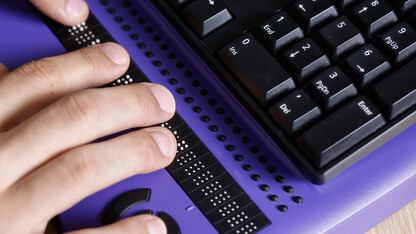 A person uses a computer with braille display and a computer keyboard.