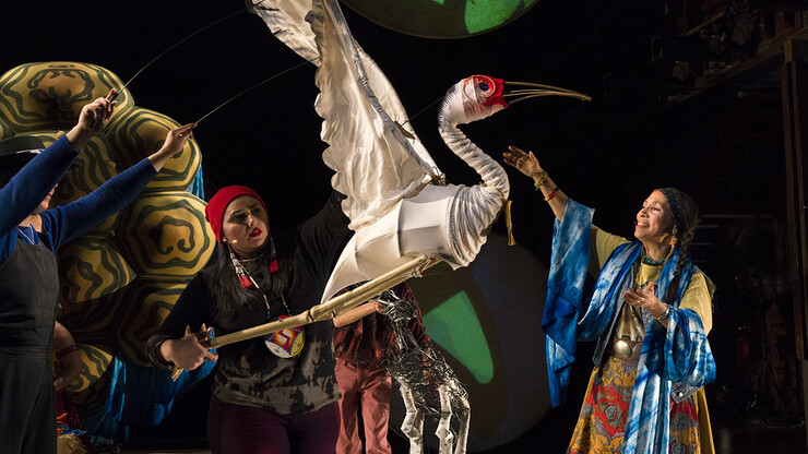 Two puppeteers control a crane puppet next to woman in colorful garments