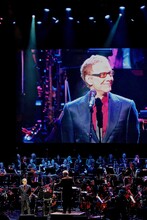 Danny Elfman stands in front of an orchestra with big screen behind