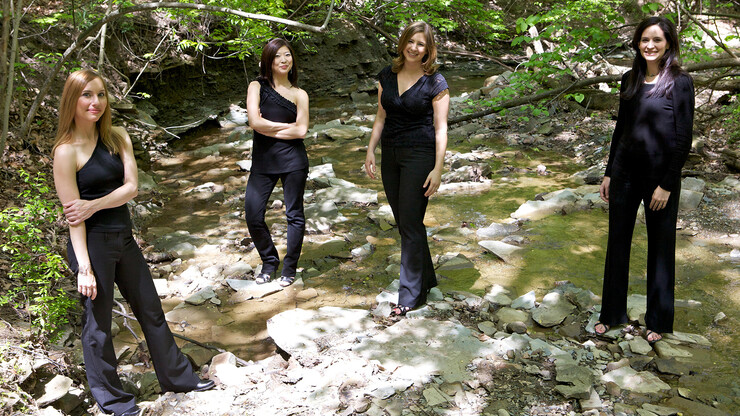 Band photo of the Omni Quartet, four women dressed in black, standing near a creek