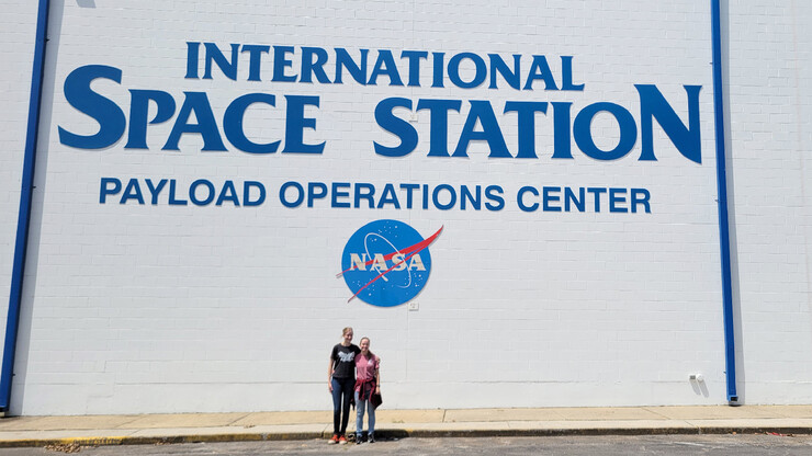 Rachael Wagner and Victoria Nelson stand in front of a NASA building that says "International Space Station Payload Operations Center."