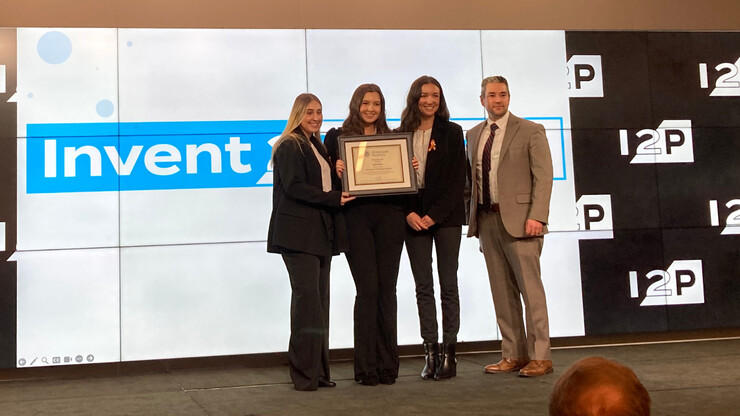 Three young women hold up a framed award next to a man, in front of a screen that says "Invent2Prevent."