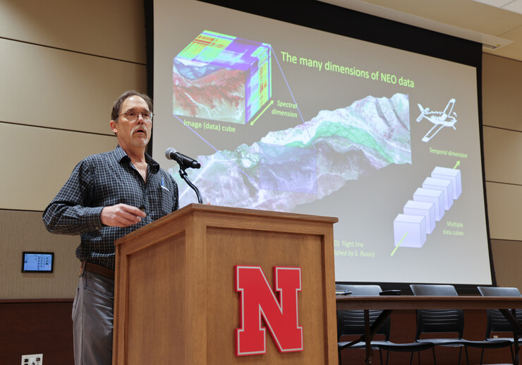 A man stands behind a lectern with a red Nebraska N on it, a screen with "The many dimensions of NEO data" in the background.