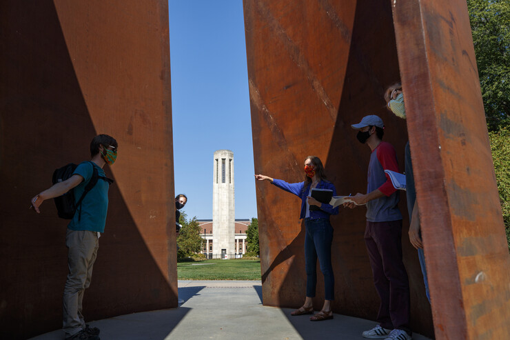 Three male students and a female instructor, wearing face masks, stand inside the "Greenpoint" sculpture, with Mueller Tower in the background.