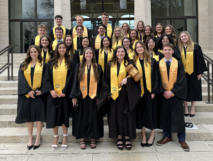 Twenty-eight college students in black robes with gold sashes pose on the staircase east of Sheldon Museum of Art.